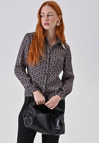 Donna con lunghi capelli rossi che indossa una camicia nera con un motivo a bottoni e pantaloni neri, mentre tiene una borsa in pelle nera a tracolla.