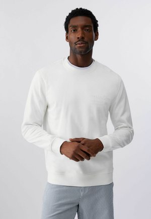 Moški, ki nosi belo majico z dolgimi rokavi in svetlo modre hlače s črtami, stoji s sklenjenimi rokami pred enobarvnim ozadjem.