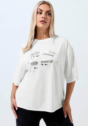 Lyshåret kvinde iført en løs hvid t-shirt med sort og sølvfarvet tekst, der siger "JEG LYTTER IKKE, MEN FORTSÆT" mod en ensartet lys baggrund.