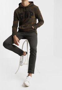 Sweat à capuche à motif camouflage avec un grand logo noir "GAP", associé à un pantalon gris foncé et des baskets blanches assis sur un tabouret.