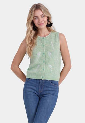Blonde vrouw met zwarte cirkelvormige oorbellen, mintgroene mouwloze top met knopen en wit bloemenpatroon, en blauwe jeans, glimlachend met handen achter haar rug.