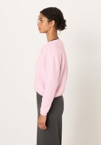 Žena s kudrnatými vlasy sepnutými dozadu má na sobě světle růžový chlupatý svetr a tmavě šedou sukni, stojící bokem proti jednobílému pozadí.