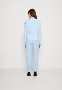 Jasnoniebieski dopasowany garnitur z taliowanym żakietem i zwężanymi spodniami. Materiał wydaje się gładki, bez widocznych wzorów. Noszony z czarnymi trampkami.