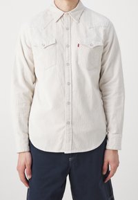 Ljust beige kordflorsskjorta med långt ärm, två bröstfickor och knapplist fram. Har spetsig krage och subtil ribbad textur.