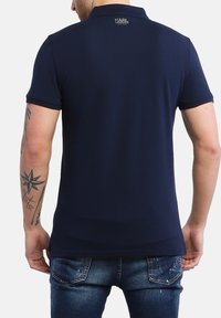 Navyblaues Poloshirt aus glattem Stoff, mit kurzen Ärmeln, einem Kragen und einem Logo auf der Rückseite. Getragen mit blauen Jeans.
