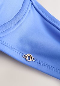 Gros plan sur une bonnette de soutien-gorge en tissu bleu avec une couture en zigzag et un petit ornement métallique circulaire argenté sur la bande.