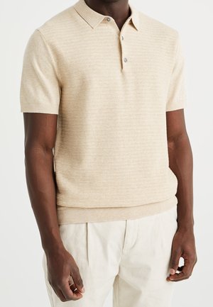 Polo beige à manches courtes avec texture côtelée, trois boutons, col plat et ourlet ajusté. Porté avec un pantalon de couleur claire.