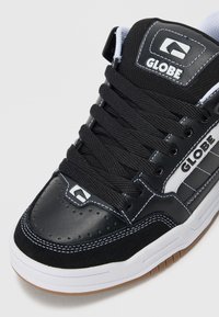 Sneaker Globe in bianco e nero con spesse stringhe nere, cuciture bianche e suola in gomma marrone, mostrata da un angolo ravvicinato frontale.