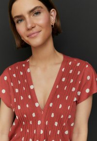 Robe rouge à manches courtes avec des pois blancs, un décolleté en V profond et des plis verticaux. Tissu doux avec une coupe décontractée.
