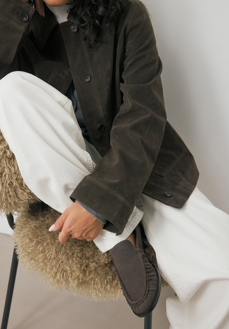 Brown suede jacket with black buttons, paired with white wide-leg pants. A hand rests on a textured cream chair, showcasing dark slip-on shoes.