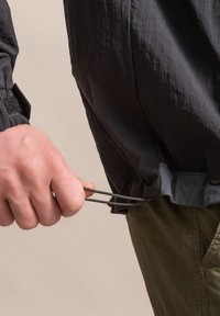 Zwarte lichtgewicht jas met een trekkoord aan de onderkant, featuring elastische manchetten en een gladde textuur. Gecombineerd met olijfgroene broek.