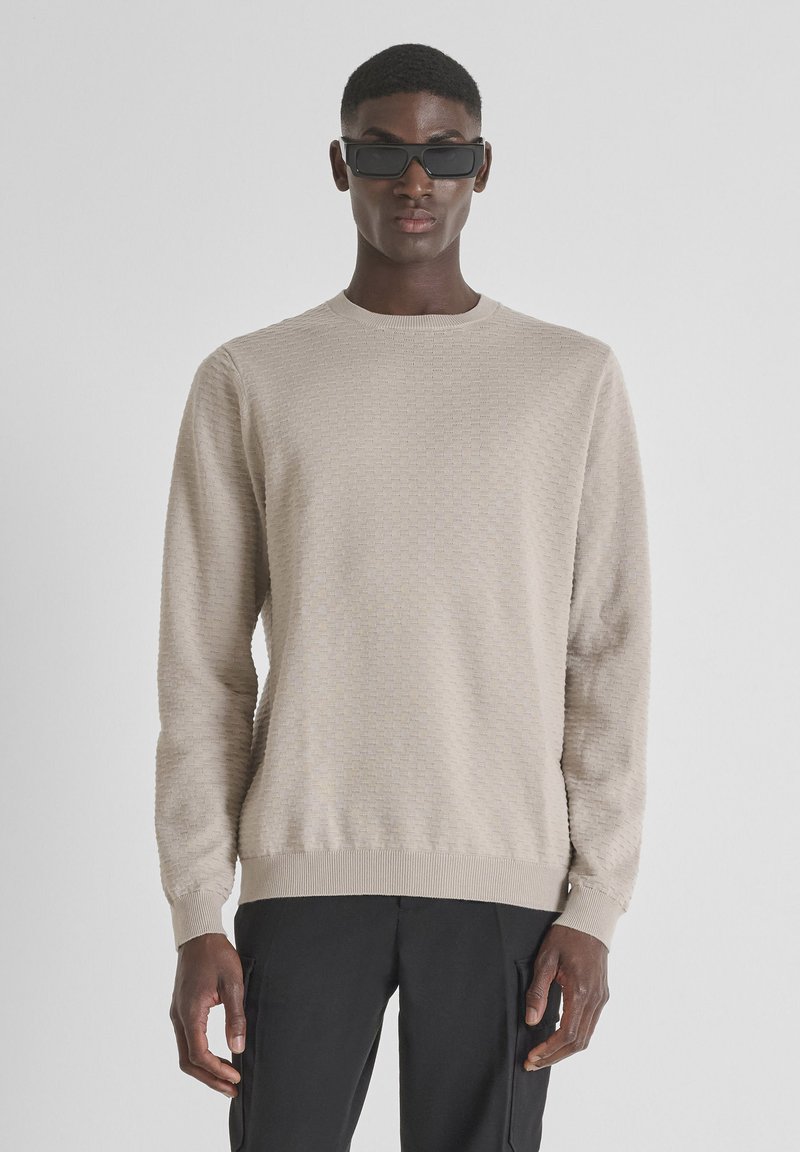 Maglione beige con texture, colletto rotondo e polsini a costine, indossato con occhiali da sole neri. Presenta un motivo geometrico sottile.
