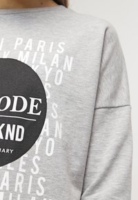 Sudadera gris con un gran emblema circular negro y texto blanco que incluye nombres de ciudades, confeccionada en una tela suave y con un corte relajado.