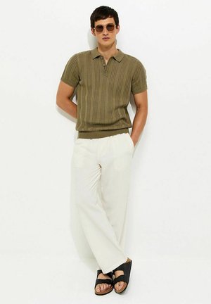 Homme portant un polo knit à manches courtes vert olive, pantalon blanc à jambes évasées, sandales noires et lunettes de soleil rondes, appuyé contre un mur blanc.