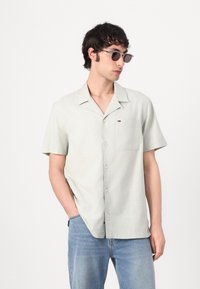 Kurzärmliges, hellgrünes Hemd mit Knopfleiste und flachem Kragen, Taschen auf der Vorderseite und subtiler Textur; kombiniert mit hellblauen Jeans.