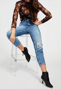 Haut en maille noir à motifs floraux avec des manches longues, associé à un jean en denim bleu clair déchiré. Bottines noires à talons pointus avec un détail de zip.