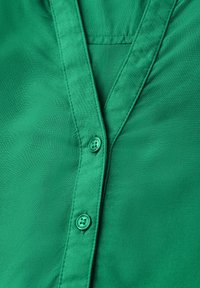 Camisa verde de botonadura frontal con una textura suave, cuello tipo notch y un único detalle de botón a juego en el frontal.