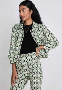 Grüner und schwarzer, gemusterter Blazer mit Drei-Knopf-Verschluss, kombiniert mit passenden, hoch taillierten Hosen, die geometrische Motive aufweisen.