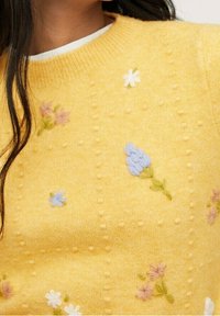 Pull en tricot jaune avec des fleurs brodées en rose, blanc et bleu, orné de petits points en relief pour la texture et doté d'un col rond.