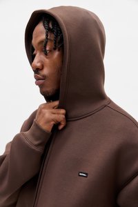 Felpa marrone con cappuccio e finitura testurizzata, caratterizzata da polsini a coste e una piccola toppa con logo nero sul davanti. Il modello si sistema il cappuccio.