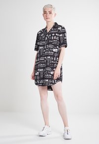 Abito a maniche corte di colore nero con stampe grafiche bianche di loghi e testo. Tessuto leggero con una vestibilità comoda e lunghezza fino a metà coscia.