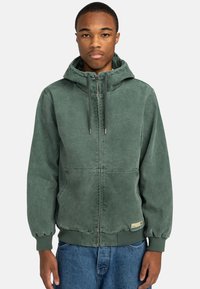 Sudadera verde con cremallera en el frente, capucha ajustable con cordón, puños y dobladillo acanalados, y dos bolsillos frontales. Tejido suave y texturizado.