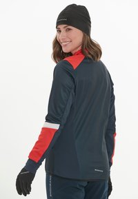 Marineblauwe sportjas met rode accenten, gestructureerde mouwen en reflecterende details. Gecombineerd met een zwarte beanie en zwarte handschoenen.