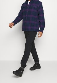 Laivastonsininen ja violetti ruudullinen flanellipaita, jossa on etutaskut. Päällä mustat housut ja kestävät mustat kengät, kävelee yksinkertaisella taustalla.