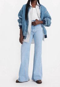 Chaqueta de mezclilla en azul medio, camisa de botones a rayas, camiseta blanca sin mangas, jeans de campana azul claro y zapatos oscuros. Textura suave en todo.