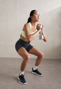 Une personne est accroupie en tenant un haltère argenté. Elle porte un soutien-gorge de sport vert clair et un short gris foncé, assortis de chaussures de sport noires.