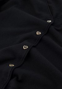 Schwarze Strickjacke mit goldenen herzförmigen Knöpfen mit dunklen Mittelpunkten auf der Vorderseite. Weiche Textur, schlichtes Design und taillierte Form.