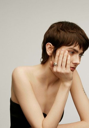 Anillo de plata llevado en la mano derecha. El sujeto tiene el cabello corto y castaño, viste una blusa negra sin tirantes frente a un fondo neutro.