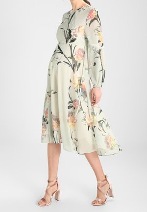 Robe fleurie en tissu vert clair avec des manches longues et une silhouette fluide. Présente des motifs floraux colorés et des détails en dentelle le long du décolleté.
