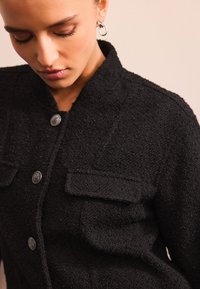 Giacca nera con texture, colletto, chiusura frontale con bottoni e due tasche sul petto; presenta bottoni metallici come dettaglio.