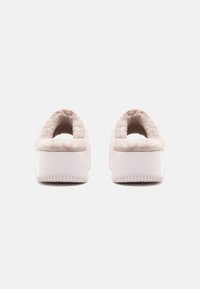 Pantuflas de felpa beiges y suaves, con una suela de goma blanca, que cuentan con un forro interior acogedor y un diseño de talón abierto para poner y quitar fácilmente.