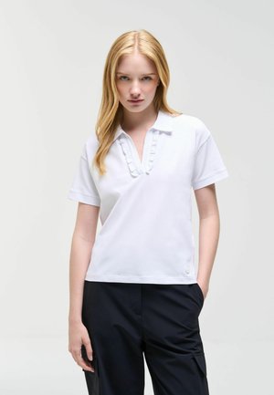 Jonge vrouw met lang blond haar draagt een wit polohemd met korte mouwen en een zwarte broek, staand met één hand in de zak.