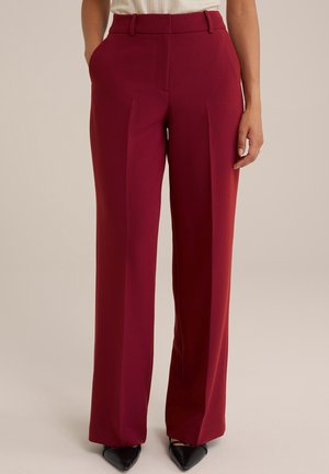 Femme portant un pantalon taille haute à jambes larges rouge foncé, associé à des chaussures noires pointues, debout devant un fond neutre.