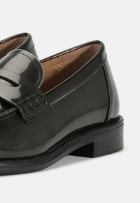 Mocasini din piele lacuită neagră cu o finisare netedă, prezentând o curea decorativă și cusături, precum și un toc de lemn low.