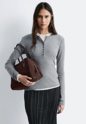 Kobieta o długich ciemnych włosach nosi szary, zapinany na guziki sweter z długim rękawem oraz czarną spódnicę, trzymając na lewej ręce brązową skórzaną torebkę.