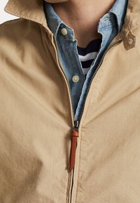 Beige bunda se zipem, s koženým táhlem, vrstvená přes světle modrou košili z chambray, ukazující kontrastní textury.