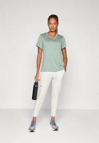 Junge Frau in hellgrünem Kurzarmshirt, weißen Hosen, grauen Sneakern, mit einer schwarzen Wasserflasche in der Hand, steht vor einem weißen Hintergrund.