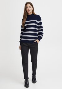 Marineblau gestreifter Pullover mit weißen horizontalen Streifen, gerippter Struktur, hohem Kragen, kombiniert mit schwarzen Hosen und eleganten Stiefeletten.