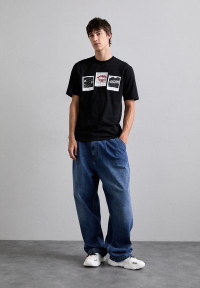 T-shirt en coton noir avec des impressions graphiques dans des cadres blancs, porté avec un jean en denim bleu ample et des baskets blanches.