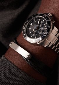 Metal watch with black dial, silver accents, and chronograph features. Worn on wrist alongside a braided black leather bracelet and silver clasp.