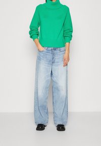 Pull vert en maille avec un col montant, accents côtelés, associé à un jean large bleu clair. Des chaussures plates noires complètent la tenue.