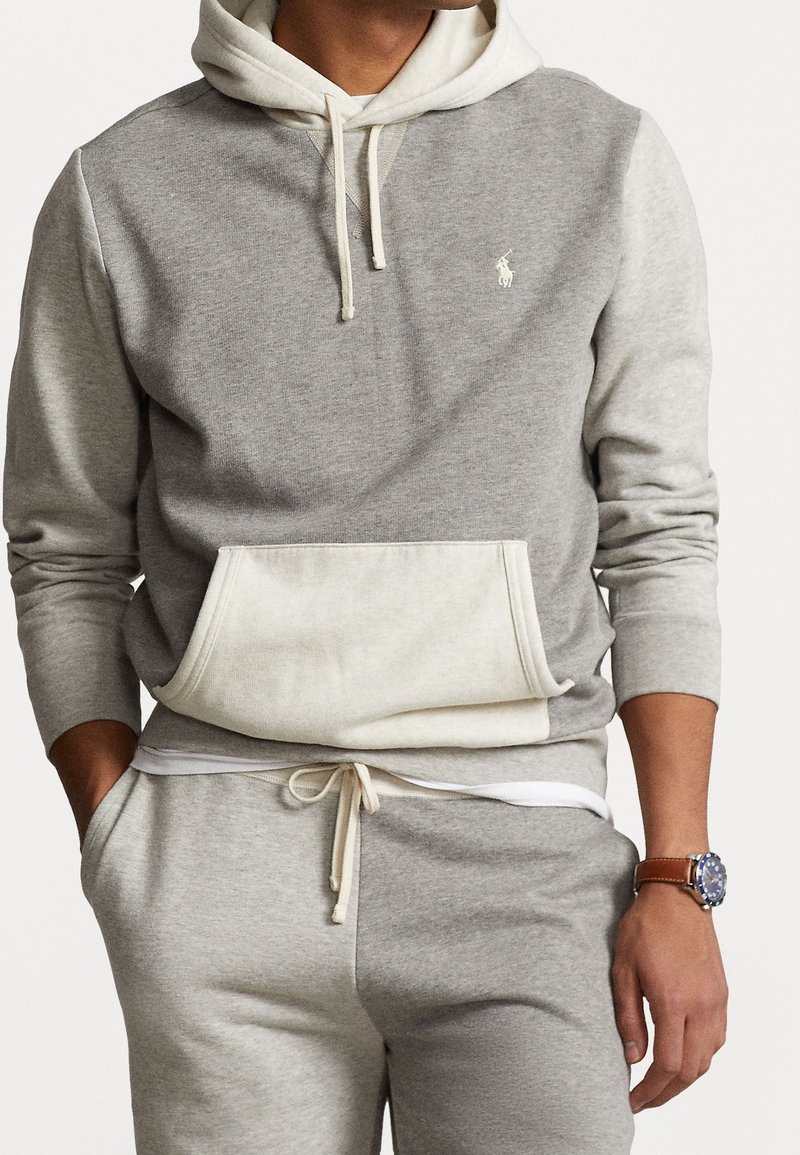 Homme portant un sweat à capuche gris et blanc cassé avec un pantalon de survêtement assorti, et une montre à bracelet en cuir marron au poignet gauche.