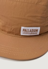 Brūna auduma cepure ar izliekto brim un balto taisnstūra plāksteri, uz kura priekšpuses ir izšūts teksts "Palladium L'Originale Depuis 1947".