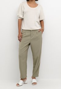 Kobieta w jasnobeżowej bluzce z krótkim rękawem, oliwkowych, wysokich na talii spodni zwężających się ku dołowi, białych sandałach na obcasie oraz złotej bransoletce.