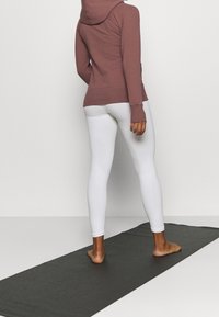 Hættebluse i mauve med tommelhuller, parret med hvide tætsiddende leggings med teksturerede mønstre. Modellen står på et sort yogamatte.
