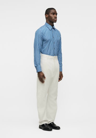 Άνδρας όρθιος με κατεύθυνση προς τα δεξιά, φορά μπλε και λευκό ριγέ μακρυμάνικο πουκάμισο, λευκό παντελόνι με ψηλή μέση και μαύρα επίσημα παπούτσια.